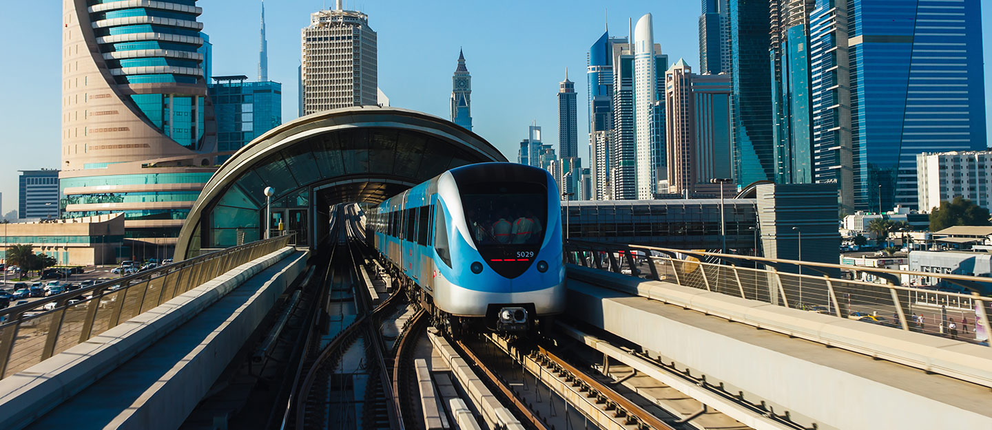 A modern train approaches a station in a city with tall skyscrapers and a clear blue sky.